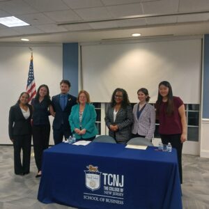 HSS Students, Senator Linda Greenstein, and Assemblywoman Verlina Reynolds-Jackson posed and smiling at the camera behind a table with a table cloth emblazoned with the TCNJ School of Business Logo.