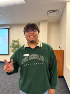 Angel Olivares in a green crewneck with "Los Angeles" emblazoned on the chest, standing for the camera holding up a peace sign in what might be a corner of the Intercultural Center.