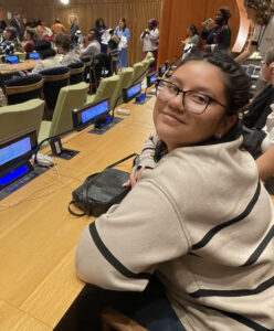 Gianella Bautista in a beige sweater with thin black horizontal stripes sitting in a lecture hall, leaning against the tabletop looking at the camera facing her left side.