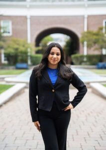 Leila Garcia Barahona posed with one hand on her hip in front of the TCNJ Science Complex fountain, dressed in a black suit with a bright blue shirt underneath.